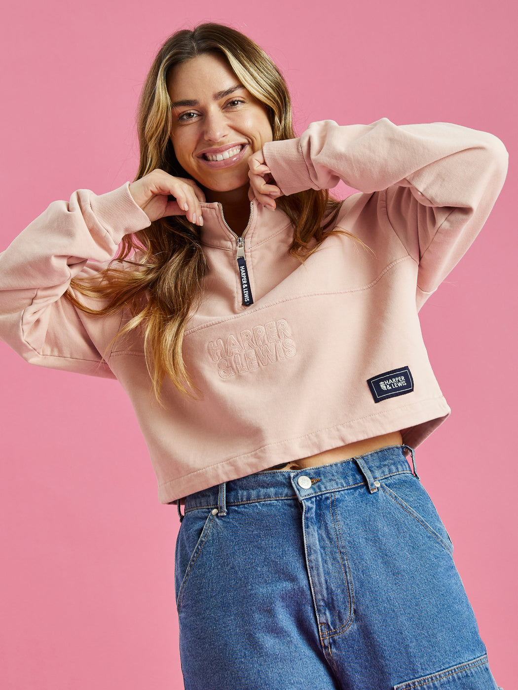 A female model wearing a dusty pink, oversized cropped 1/4 zip sweater with Harper & Lewis tonal spellout, paired with blue jeans.