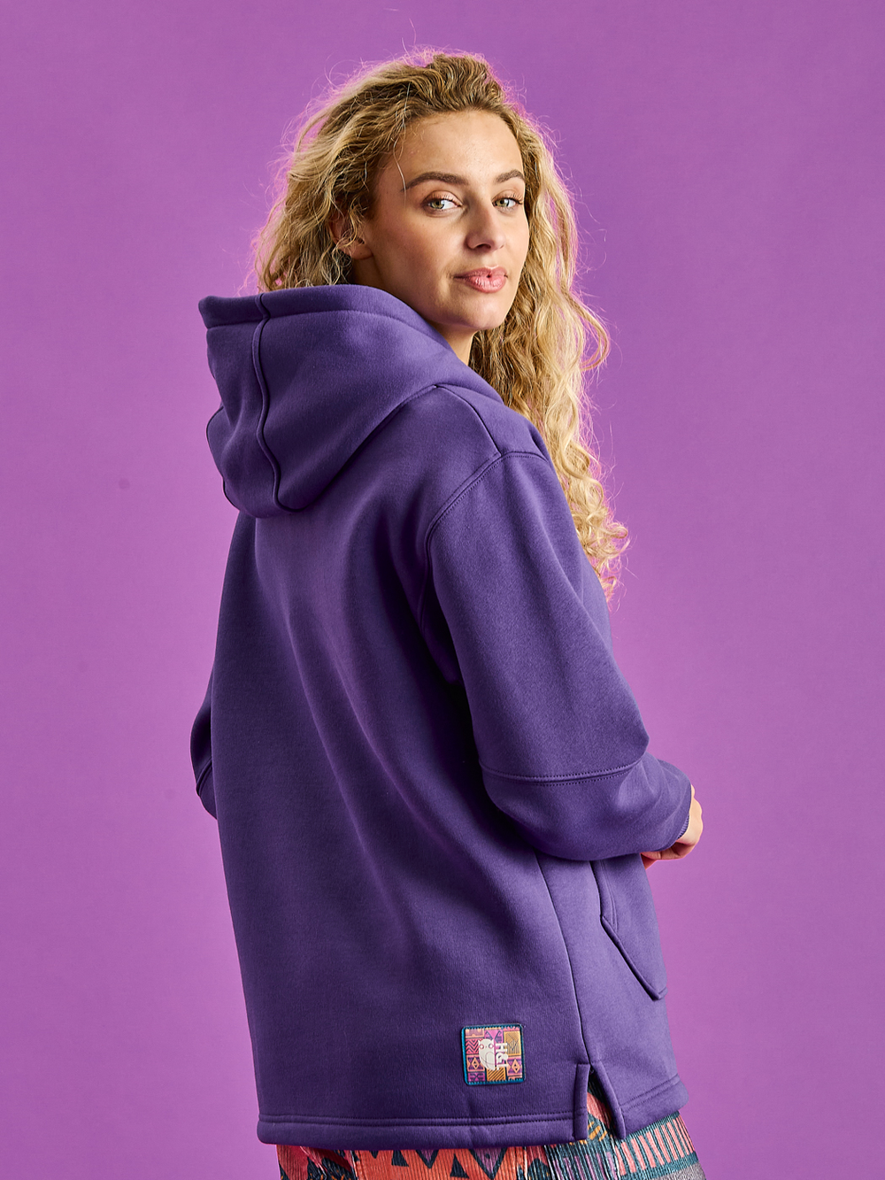 Female Model wears the indiana Hooded sweater in purple showing the branded woven badge on the hem.