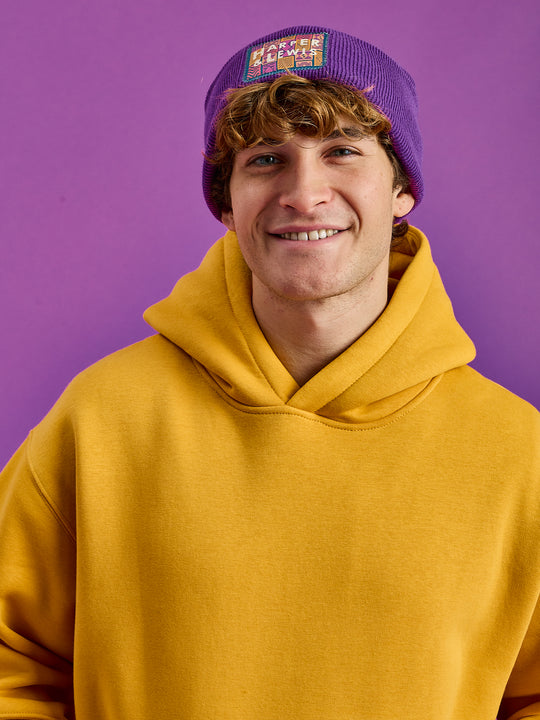 Male Model wears a Yellow oversized unisex hoodie with a purple beanie hat with matching badges.