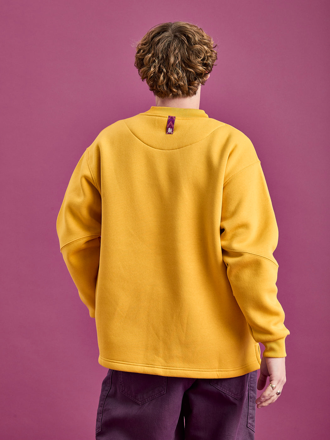 Back shot of the male model wearing the Winnie sweater in Mustard yellow, showing the branded woven tab at the back of the neck which can be used as a hanging loop.
