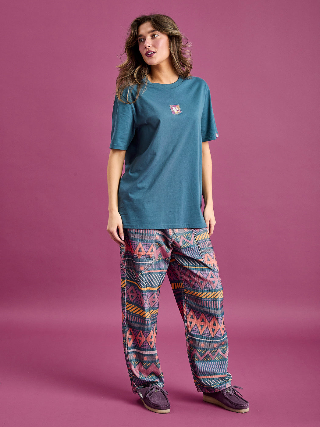Female model wears the Neva T-shirt in a Petrol Blue colour, featuring a front badge detail.