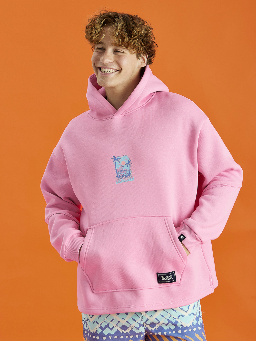 Male Model wearing the Cruz hoodie in Pink, this is adorned with a Tropical Graphic on the back and a Harper & Lewis emblem on the front, a nod to our vintage-inspired roots.