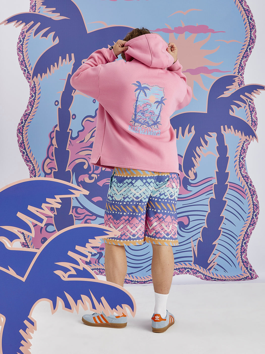 Male Model wearing the Cruz Hoodie in Pink, this is showing the back print on the, which is tropical inspired with a wave and palm tree graphic.